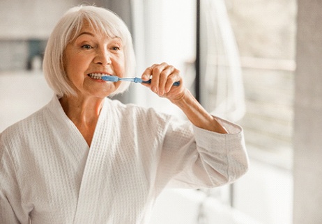 An older woman wearing a white bathrobe and brushing your dental implants in her bathroom